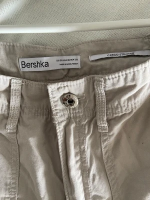 Beige cargobyxor från Bershka - Snygga beige cargobyxor från Bershka med raka ben och flera fickor, inklusive sidofickor med lock och knapp. Byxorna har justerbart midjeband med spänne och är tillverkade i ett slitstarkt bomullsmaterial. 