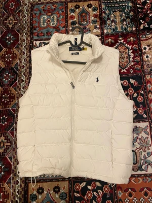 Vit dunväst från Polo Ralph Lauren - Snygg vit dunväst från Polo Ralph Lauren med hög krage och dragkedja framtill. Västen har det klassiska Polo-logotypen broderad på bröstet och quiltad design. Perfekt för lager-på-lager och riktigt clean look.