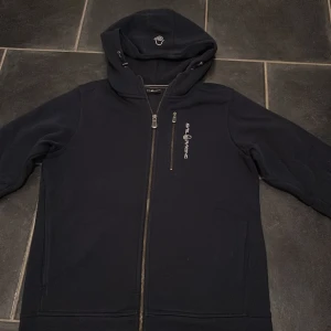 Mörkblå hoodie från Sail Racing - Snygg mörkblå hoodie från Sail Racing i storlek 170,med dragkedja framtill och logga på bröstet. Huva med broderad logga och en extra ficka med dragkedja på bröstet. Använd bara 2 gånger så i väldigt bra skick 