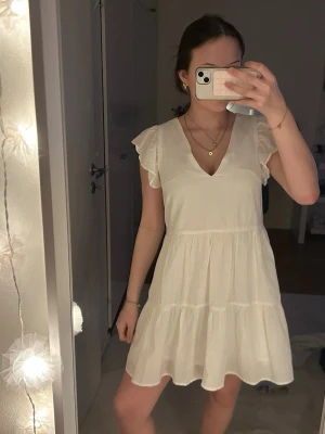Vit kortklänning med volangärm - Söt vit kortklänning med v-ringning och volangärmar. Klänningen har en luftig passform och är sydd i flera lager för en flowy känsla. Perfekt för varma dagar och enkel att styla med smycken. Märke: H&M.