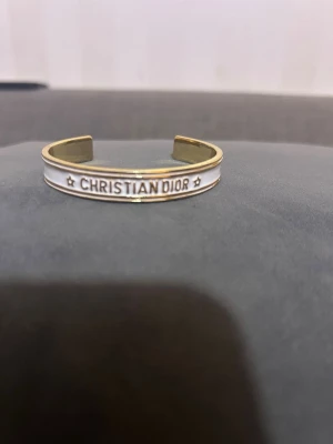 Vitt och guldigt Dior armband - Snyggt armband från Christian Dior i guldtonad metall med vit emalj och stjärndetaljer. Öppen design som gör det enkelt att ta på och av. Märkets logga är tydligt ingraverad framtill för en exklusiv känsla.