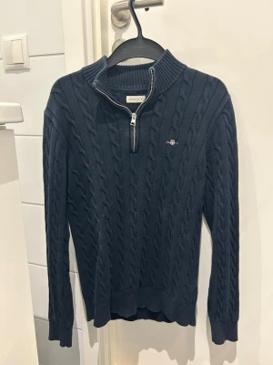Mörkblå kabelstickad Gant tröja - Mörkblå kabelstickad tröja från Gant med halv dragkedja och hög krage. Tröjan har ribbade muddar och nederkant samt en liten broderad logga på bröstet. Perfekt för dig som gillar klassisk och stilren design. 