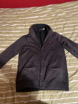 Cardigan från Ralph Lauren - Snygg Cardigan från Ralph Lauren med sjalkrage och knäppning framtill. Koftan har den klassiska röda logotypen broderad på. Perfekt för lager på lager, nu inför våren och en stilren look. Cardiganen är använd ett par gånger då den är rätt stor på mig, väldigt lätt att styla också. Storleken är S och priset jag frågar efter är 400. Om du vill ha fler bilder eller så är det bara att hojta till!