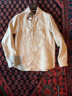 Beige linneskjorta från Oscar Jacobson och neil barret t shirt - Stilren beige skjorta från Oscar Jacobson i linne med klassisk krage, bröstfickor med knappar och långa ärmar. Perfekt för en avslappnad men ändå snygg look. T shirt också 