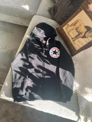 Svart hoodie med dragkedja - Unik hoodie med dragkedja och stor vit All Star-logga på både bröstet och ryggen. Klassisk huva och känguruficka framtill. Perfekt för en avslappnad streetstyle-look. Storlek 176. 