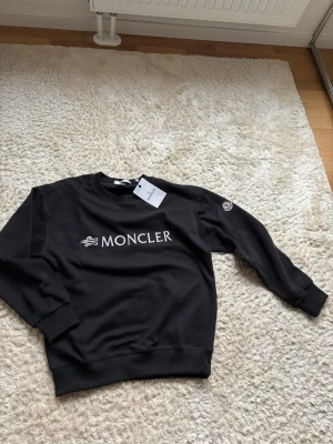 Svart sweatshirt från Moncler - Stilren svart sweatshirt från Moncler med vit logotyp på bröstet och Moncler-märke på ärmen. Tröjan har rund halsringning, långa ärmar och ribbade muddar. Perfekt för en avslappnad och trendig look.