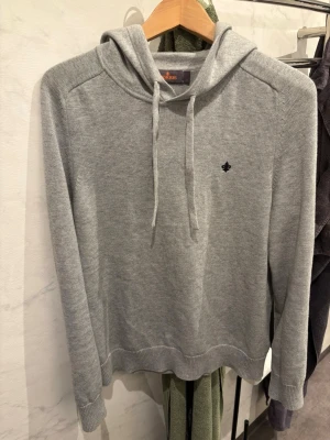 Morris Hoodie - Grå, finstickad huvtröja från Morris. Tröjan har huva med snörning samt ribbstickade muddar vid ärm- och plaggslut och broderad logotyp på bröstet. Tillverkad i en bomull- och merinoullblandning. Nypris 2000 finns ej i lager längre