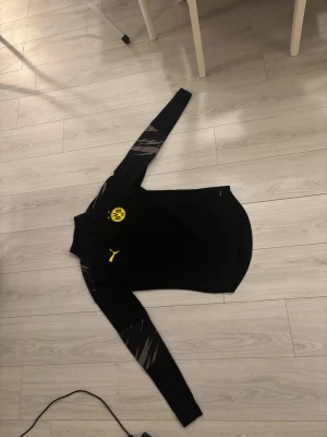 BVB x Puma svart träningsset - Säljer ett svart träningsset från Puma och Borussia Dortmund (BVB). Setet består av en långärmad tröja och matchande byxor, båda med gula Puma-loggor och BVB-emblem. Materialet är troligtvis polyester, perfekt för träning eller chill. Snygga detaljer på ärmarna.
