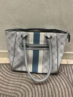 Grå handväska med mönster och ränder - Snygg grå handväska med vita och blå vertikala ränder framtill. Väskan har ett diskret mönster, dubbla handtag och en dragkedja upptill. Invändigt finns flera fack, inklusive ett med dragkedja. Perfekt för dig som gillar stilrena detaljer.