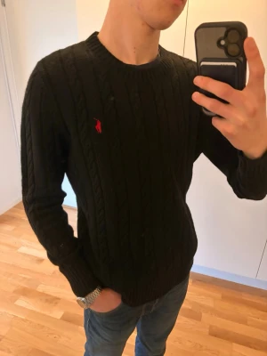 Svart kabelstickad tröja Polo Ralph Lauren - Kabelstickad svart tröja från Polo Ralph Lauren med röd broderad logga på bröstet. Rund halsringning och ribbade muddar vid ärmslut och nederkant. Klassisk och stilren modell som passar till jeans eller chinos.