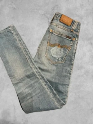 Ljusblå slitna jeans från nudie jeans co - Säljer ett par ljusblå jeans från Replay med slitningar och patchdetalj på bakfickan. Jeansen har klassisk femficksmodell, raka ben och snygga slitningar framtill och baktill. Perfekta för en avslappnad och trendig look.