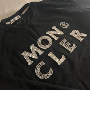 Svart Moncler sweatshirt med mönstrad logga - Snygg svart sweatshirt från Moncler med stora bokstäver och logga framtill i ett coolt mönstrat tryck. Tröjan har rund halsringning och långa ärmar. Perfekt för dig som gillar streetwear och vill ha något som sticker ut. Litet hål i armhålan syns inte när man har på sig de därav priset 