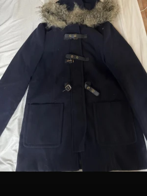 Marinblå duffelrock med fuskpäls - Snygg marinblå duffelrock med huva kantad av fuskpäls. Klassiska träknappar med läderöglor och stora framfickor. Rocken har rak passform och är perfekt för kyliga dagar. Materialet ser ut att vara ullblandning.