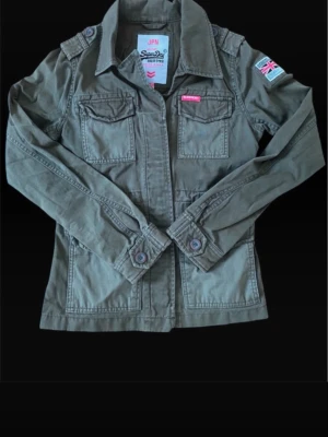 Grön vindjacka från Superdry - Stilren grön vindjacka från Superdry med två bröstfickor, axelklaffar och patch med brittisk flagga på ärmen. Jackan har klassisk krage, knappdetaljer vid ärmslut och är tillverkad i bomull. Perfekt för dig som gillar en militärinspirerad look.