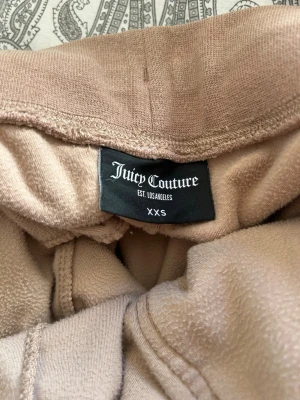 Beiga velourbyxor Juicy Couture - Säljer ett par beiga mjukisbyxor från Juicy Couture i velour. Byxorna har bred mudd i midjan, dragsko och fickor fram. Klassisk logga broderad på ena fickan. Perfekta för en chill och trendig look.
