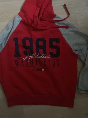 Röd och grå hoodie 1985 Washington - Snygg hoodie i rött och grått med trycket '1985 Athletics Washington USA' på bröstet. Hoodien har huva med dragsko och kontrasterande ärmar. Perfekt för en sportig och avslappnad look.