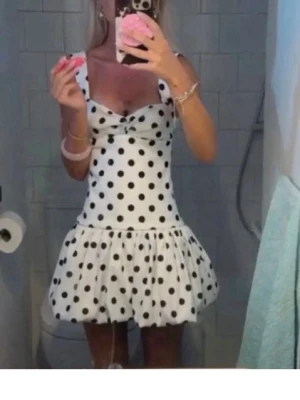 Vit prickig kortklänning - Supersöt vit kortklänning med svarta prickar och breda axelband. Klänningen har en markerad midja och en volangig kjol som ger en lekfull look. Perfekt för dig som vill sticka ut med ett klassiskt men trendigt mönster.