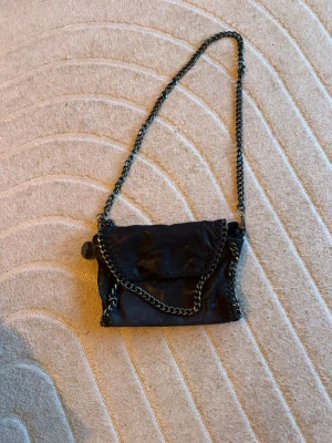 Svart kedjehandväska från Stella McCartney - Cool svart handväska från Stella McCartney med kedjedetaljer runt kanterna och som axelrem. Väskan har en mjuk, glansig yta och lock med magnetknäppning. Perfekt för dig som gillar edgy och trendig stil.