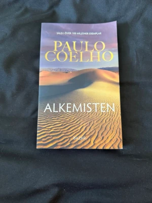 Alkemisten av Paulo Coelho - En svensk pocketutgåva av Paulo Coelhos roman 'Alkemisten' med ett färgstarkt omslag som visar en öken i gult och orange. Boken har tydlig titel och författarnamn i stora bokstäver på framsidan.
