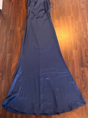 Marinblå långklänning i satin - Elegant marinblå långklänning i glansigt satinliknande material. Klänningen har smala axelband och en v-ringad urringning. Perfekt för speciella tillfällen när du vill känna dig extra fin.