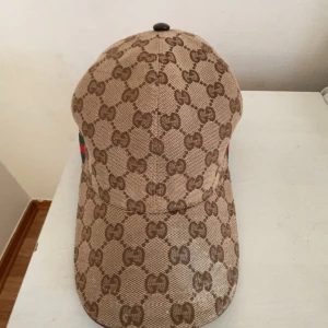 Beige keps med GG-mönster från Gucci - Säljer en beige keps från Gucci med det klassiska GG-monogrammönstret över hela kepsen. Kepsen har böjd skärm, justerbart spänne bak och är tillverkad i ett slitstarkt tygmaterial. Perfekt för dig som vill ha en ikonisk och trendig accessoar.