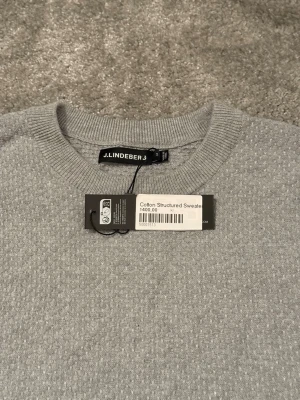 Ljusgrå stickad tröja J.Lindeberg - Snygg ljusgrå stickad tröja från J.Lindeberg i bomull med diskret strukturerat mönster. Tröjan har rund halsringning och långa ärmar, perfekt för en stilren och enkel look. Helt ny med prislapp, köpt för 1400