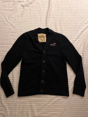 Svart kofta från Hollister L - Kofta från Hollister i svart med långa ärmar och knappar framtill. Klassisk design med broderad logga i rött och vitt på bröstet. Perfekt för lager på lager och enkel att matcha till olika outfits. Storlek L skriv om ni undrar nått fraktar direkt😊