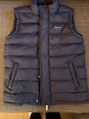 Mörkblå dunväst från GANT - Snygg mörkblå dunväst från GANT med röd insida och diskret logga på bröstet. Västen har två snedställda fickor framtill och stängs med dragkedja och tryckknappar. Perfekt för lager-på-lager och håller dig varm med sin vadderade design. Västen är snålt använd och i ett bra skick. 