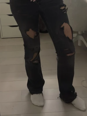 Gina tricot Low waist jeans - Säljer ett par svarta jeans med stora slitningar och hål. Boot cut, low waist, och fickor med knappar på baksidan av jeansen. Perfekta för dig som vill sticka ut med en trendig look.
