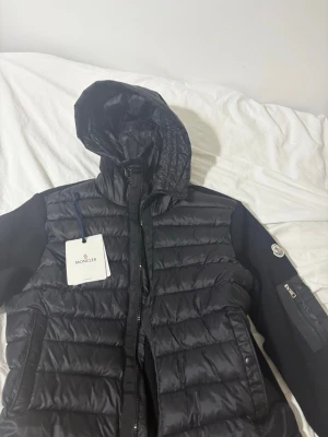 Moncler cardigan - Snygg svart dunjacka från Moncler med quiltad design och huva. Jackan har dragkedja framtill, två sidofickor med tryckknappar och Moncler-logga på ärmen. Perfekt för kalla vinterdagar och riktigt stilren look.