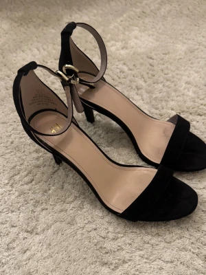 Svarta pumps med ankelrem från H&M - Stilrena svarta pumps från H&M med öppen tå och smal ankelrem med guldfärgat spänne. Klacken är smal och hög, och materialet ser ut att vara mockaimitation. Perfekta för att lyfta vilken outfit som helst.
