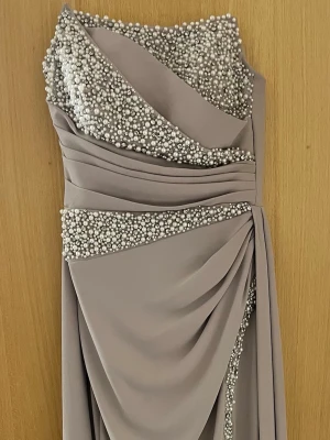 Långklänning med pärldetaljer - Elegant långklänning i ljusbeige med pärlbroderier över byst och midja. Klänningen har draperad design, axelbandslös skärning och svepande tyg. Perfekt för speciella tillfällen där du vill sticka ut med exklusiva detaljer.