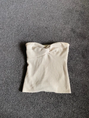 Beige ribbad bandeau tubtopp - Säljer en beige ribbad bandeau tubtopp med twistad detalj framtill. Toppen är axelbandslös och har en mjuk, stretchig passform som sitter snyggt på kroppen. Perfekt att styla till jeans eller kjol för en clean look🥂💕