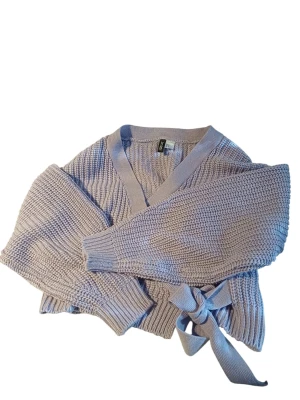 Ljuslila omlottkofta från H&M - Supersöt ljuslila stickad omlottkofta från H&M med breda ribbade muddar och knytband i midjan. Koftan har en djup V-ringning och är gjord i mjukt, grovstickat material. 