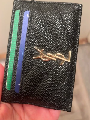 Svart YSL korthållare i läder - Snygg svart korthållare från Saint Laurent i strukturerat läder med guldfärgad YSL-logga framtill. Smidig och stilren design med plats för flera kort. Perfekt accessoar för dig som vill ha något både lyxigt och praktiskt.