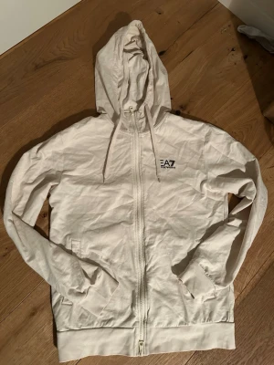 EA7 zip hoodie - Beige tunn EA7 zip hoodie i storlek S. Hoodien är perfekt för träning eftersom den är lite tunnare eller bara till en vanlig outfit. Den är sparsamt använd och i väldigt fint skick. Skriv om du har några frågor!