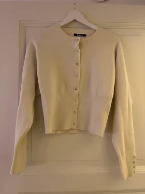 Beige cardigan från Gina Tricot - Stilren beige cardigan från Gina Tricot med rund halsringning och silvriga knappar framtill. Tröjan är ribbstickad vid hals, knappslå och ärmslut. Perfekt att slänga över en topp för en enkel men snygg look.