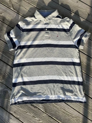 Vintage Randig piké Tommy Hilfiger 2000s - Snygg pikétröja från Tommy Hilfiger med breda marinblå, vita och grå ränder. Klassisk krage, korta ärmar och liten logga på bröstet. Perfekt till jeans eller shorts för en avslappnad stil.