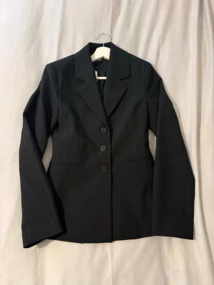 Svart kavaj från Gina Tricot - Stilren svart kavaj från Gina Tricot med tre knappar framtill och klassisk krage. Figurnära passform och diskreta fickor. Perfekt för en trendig och snygg look. Materialet är mjukt och följsamt, vilket gör den bekväm att bära hela dagen.