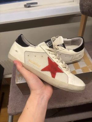 Golden Goose  - Golden Goose sneakers i vitt läder och mesh med en ikonisk röd stjärna på sidan och svart detalj vid hälen. Skorna har vita snören, GGDB-märke på plösen och guldfärgad text på sidan. Snygg streetstil med vintage vibe. Finns en liten spricka på sulan, se bild 4, annars mycket gott skick 