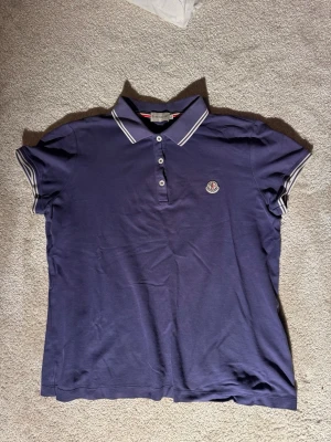 Marinblå pikétröja från Moncler - Snygg blå pikétröja från Moncler med vitrandiga detaljer på krage och ärmslut. Klassisk polokrage med tre knappar framtill och Moncler-logga på bröstet. Perfekt till jeans eller shorts för en clean look. Säljer det eftersom att jag har växt ur den!