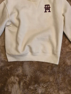 Vit sweatshirt från Tommy Hilfiger - Säljer en stilren vit sweatshirt från Tommy Hilfiger med mörkrött TH-logotypbroderi på bröstet. Tröjan har rund halsringning, ribbade muddar och är gjord i mjukt bomullsmaterial. Perfekt för en clean och avslappnad look. Nypris 1300 kr