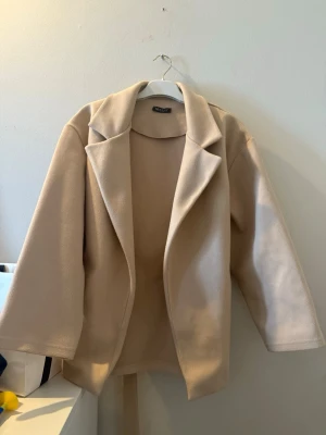 Beige kappa med knytskärp - Stilren beige kappa från Gina Tricot med bred krage och knytskärp i midjan. Kappan har en rak passform och är tillverkad i ett mjukt tyg som ger en clean och modern look. Perfekt för dig som gillar minimalistisk stil.