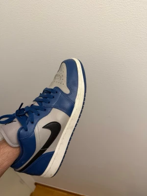 Nike Air Jordan 1 Mid blå/grå - Säljer ett par Nike Air Jordan 1 Mid sneakers i blått och grått läder med svart swoosh. Klassisk siluett med vita sulor och blå snören. Perfekta för dig som gillar streetwear och vill ha en ikonisk look.