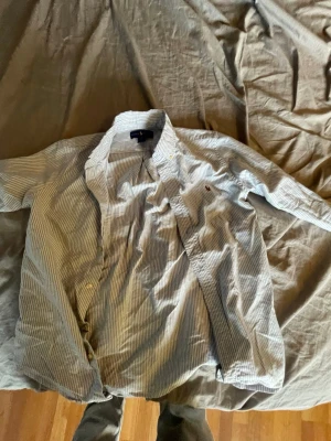 Blårandig skjorta från Polo  - Klassisk blårandig skjorta från Polo med vit botten och tunna blå ränder. Skjortan har lång ärm, knappar framtill och en liten broderad logga på bröstet. Perfekt till jeans eller chinos för en clean och stilren look.