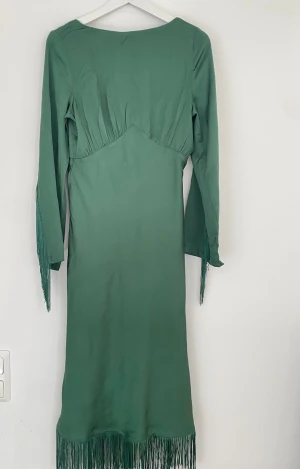 Grön långklänning med fransar - Snygg grön långklänning med långa ärmar och fransdetaljer längs ärmslut och nederkant. Klänningen har en markerad midja och är tillverkad i ett mjukt, slätt material som ger ett elegant fall. Perfekt för dig som vill sticka ut med coola detaljer.