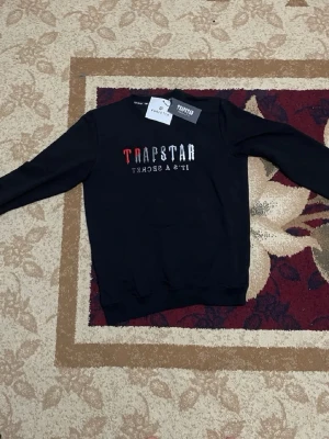 Svart sweatshirt från Trapstar - Svart sweatshirt från Trapstar med tryckt text 'Trapstar It's A Secret' i vitt och rött på bröstet. Klassisk rund halsringning och långa ärmar. Ribbstickade muddar vid ärmslut och nederkant. Perfekt för en streetwear-look.