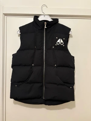 Svart dunväst från Moose Knuckles - Snygg svart dunväst från Moose Knuckles med hög krage och silverfärgad dragkedja. Västen har två framfickor med tryckknappar och det ikoniska Moose Knuckles-logotypen i vitt på bröstet. Perfekt för lager-på-lager och håller dig varm under kyliga dagar.