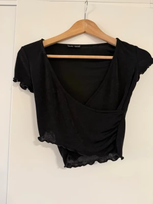 Svart omlottopp med volangkant - Snygg svart omlottopp med korta ärmar och volangkant vid ärmslut och nederkant. Toppen har en djup v-ringning och croppad passform som ger en trendig look. Perfekt att matcha med höga jeans eller kjol.