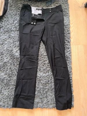 Svarta kostymbyxor - Snygga svarta kostymbyxor med raka ben och klassisk passform. Byxorna har hällor för skärp, två knappar och dragkedja i gylfen. Perfekta för en stilren och clean look. Materialet är slätt och lätt, vilket ger en dressad känsla.